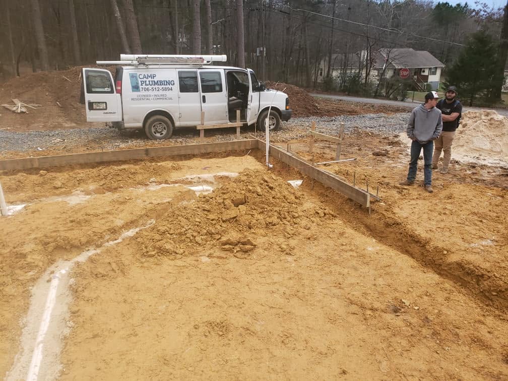 Trenched building pad with plumbing lines set and two crew members reviewing the layout